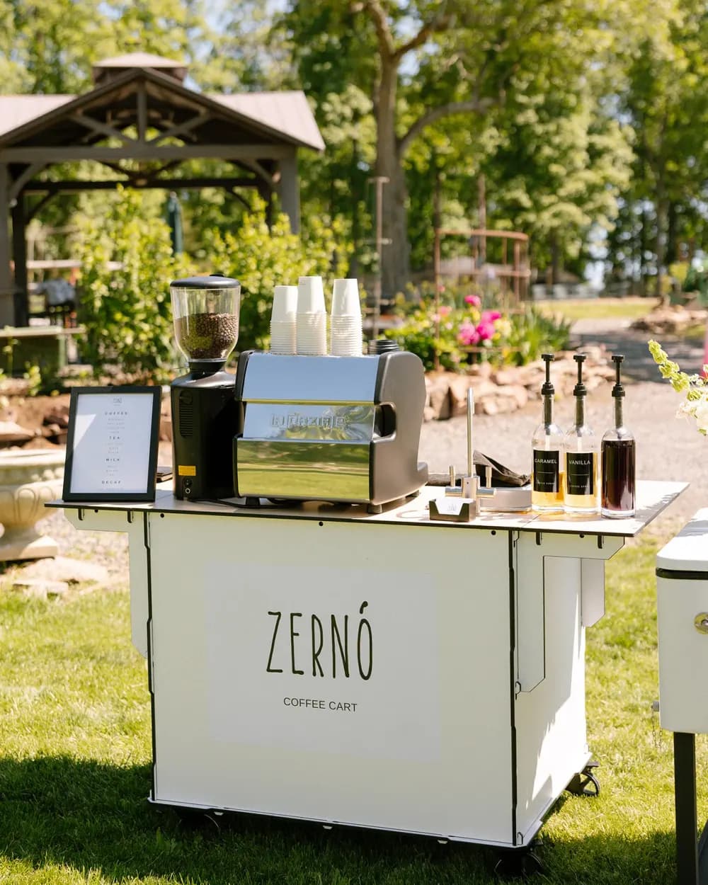 The Zerno espresso cart with grinder, espresso machine, cups and syrup bottles set up on a lawn beside a garden.