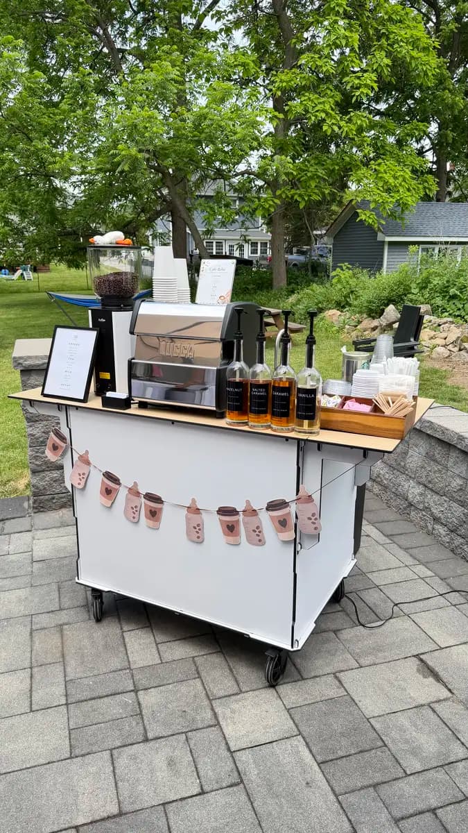 White espresso cart with cups and syrups on a paved patio beneath trees
