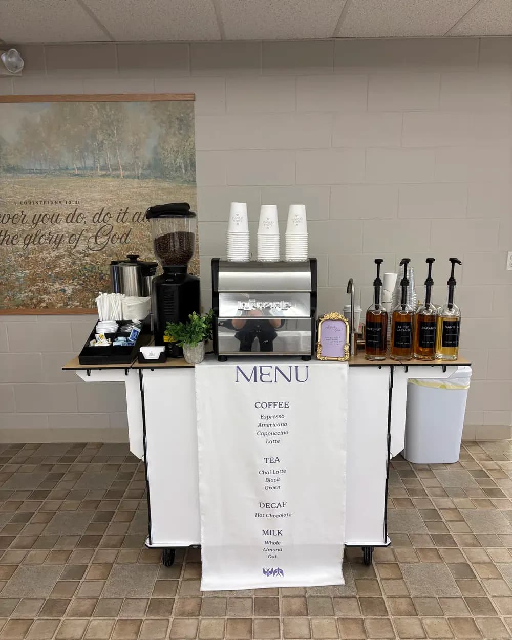 A white Zerno espresso cart with a cloth menu banner listing coffee, tea, decaf and milk options, cups stacked on the machine and four syrup bottles.